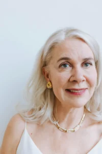 Retrato de uma mulher idosa com cabelo loiro prateado, usando joias douradas, sorrindo suavemente, em fundo branco, transmitindo elegância e experiência.