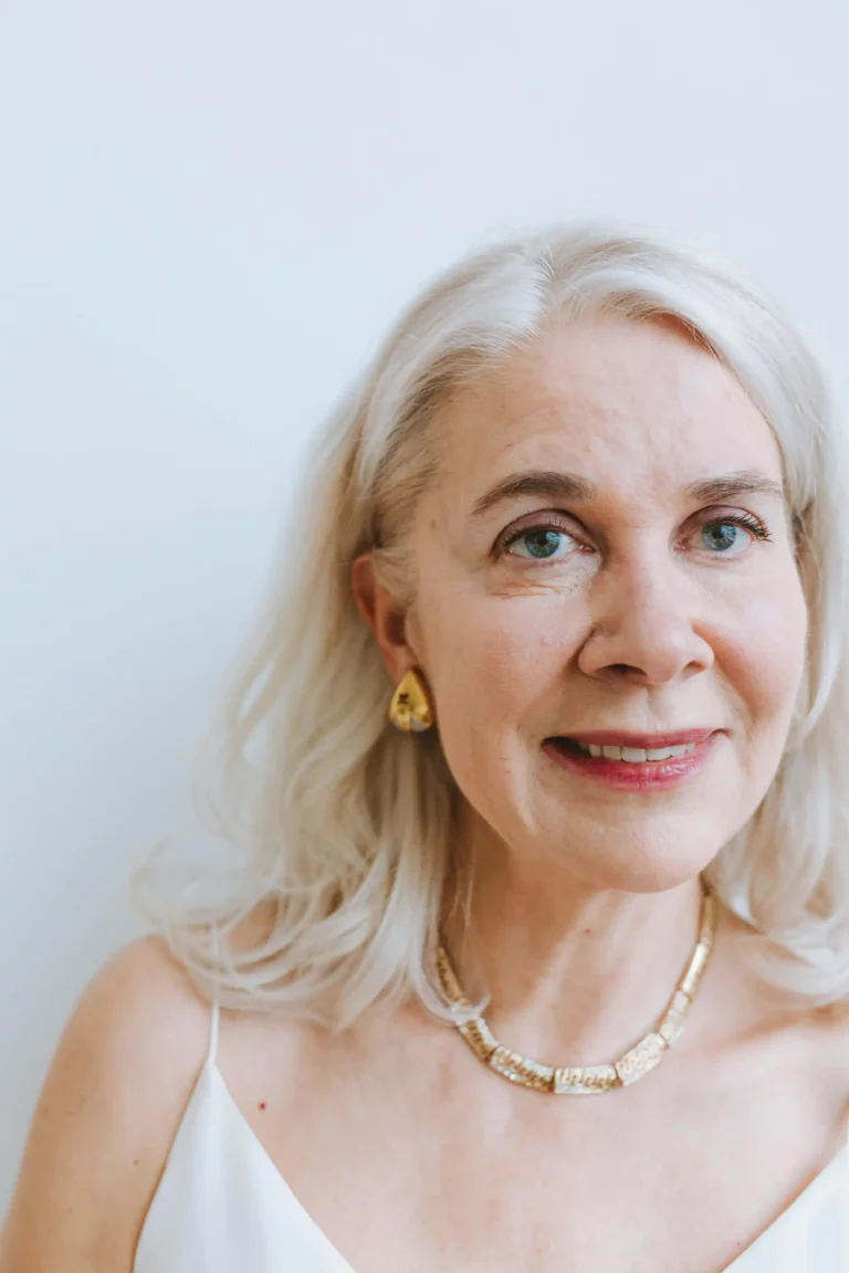 Retrato de uma mulher idosa com cabelo loiro prateado, usando joias douradas, sorrindo suavemente, em fundo branco, transmitindo elegância e experiência.