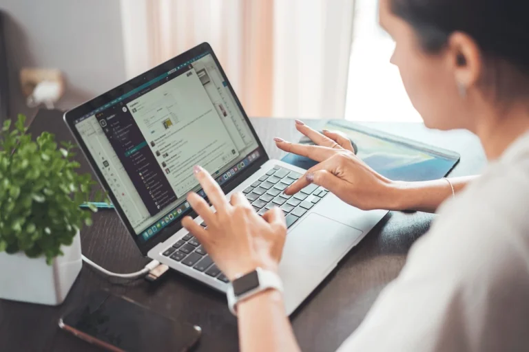 Mulher usando laptop em ambiente de trabalho com foco em produtividade digital, rodeada de objetos de escritório, incluindo uma planta.