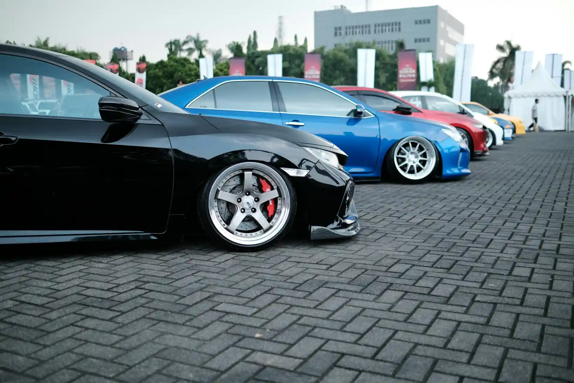 Linha de carros esportivos alinhados em uma exposição, destaque para o carro preto com detalhes em vermelho e rodas esportivas, em fundo de dia ensolarado.