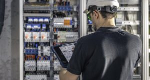 Técnico de manutenção elétrica inspecionando um quadro de distribuição de energia com um tablet em mãos, usando capacete de segurança e óculos de proteção em ambiente industrial.