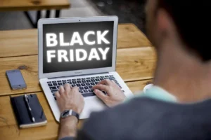 Pessoa olhando para a tela do laptop com a frase 'Black Friday', próximo a um celular e um caderno em uma mesa de madeira, promovendo ofertas especiais.