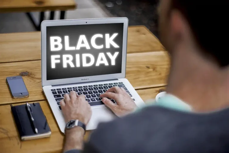 Pessoa olhando para a tela do laptop com a frase 'Black Friday', próximo a um celular e um caderno em uma mesa de madeira, promovendo ofertas especiais.