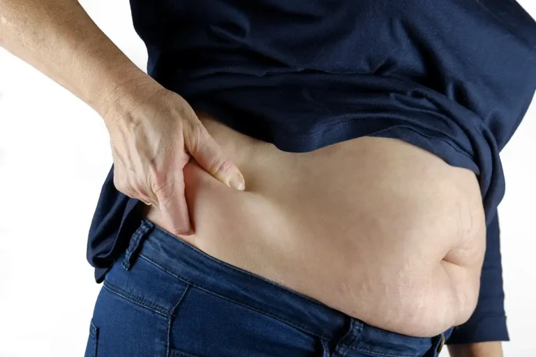 Pessoa segurando a barriga e mostrando excesso de peso na região abdominal, usando roupas casuais de cor escura e jeans.