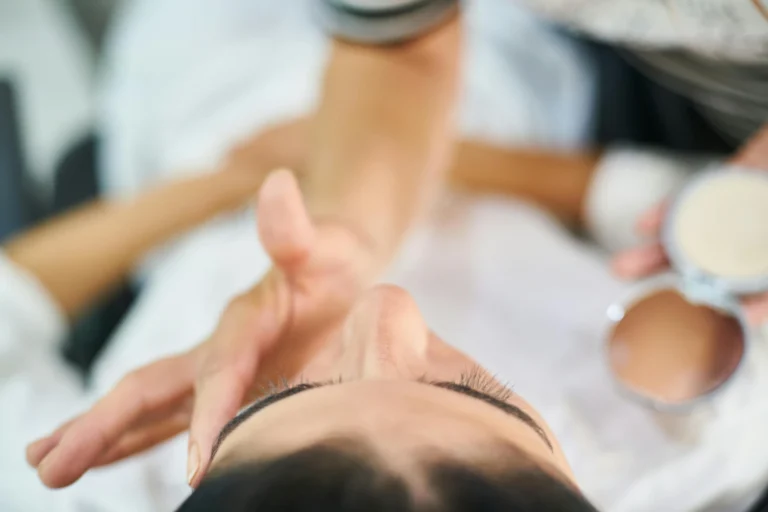 Mão de uma profissional aplicando maquiagem em uma mulher em um ambiente de beleza ou estética, foco na mão e no rosto da cliente.