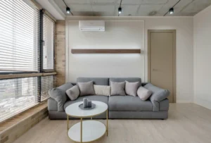 Imagem de uma sala de estar moderna com sofá cinza, almofadas, mesa de centro redonda e janelas grandes com persianas em ambiente bem iluminado.