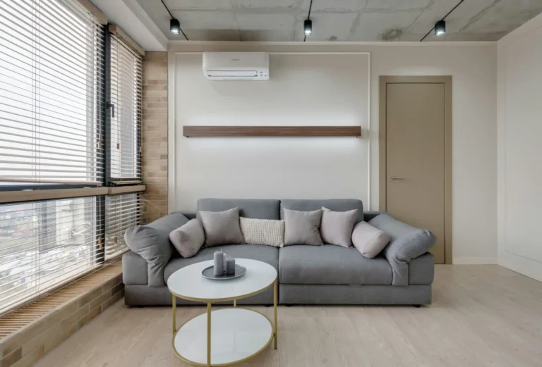 Imagem de uma sala de estar moderna com sofá cinza, almofadas, mesa de centro redonda e janelas grandes com persianas em ambiente bem iluminado.
